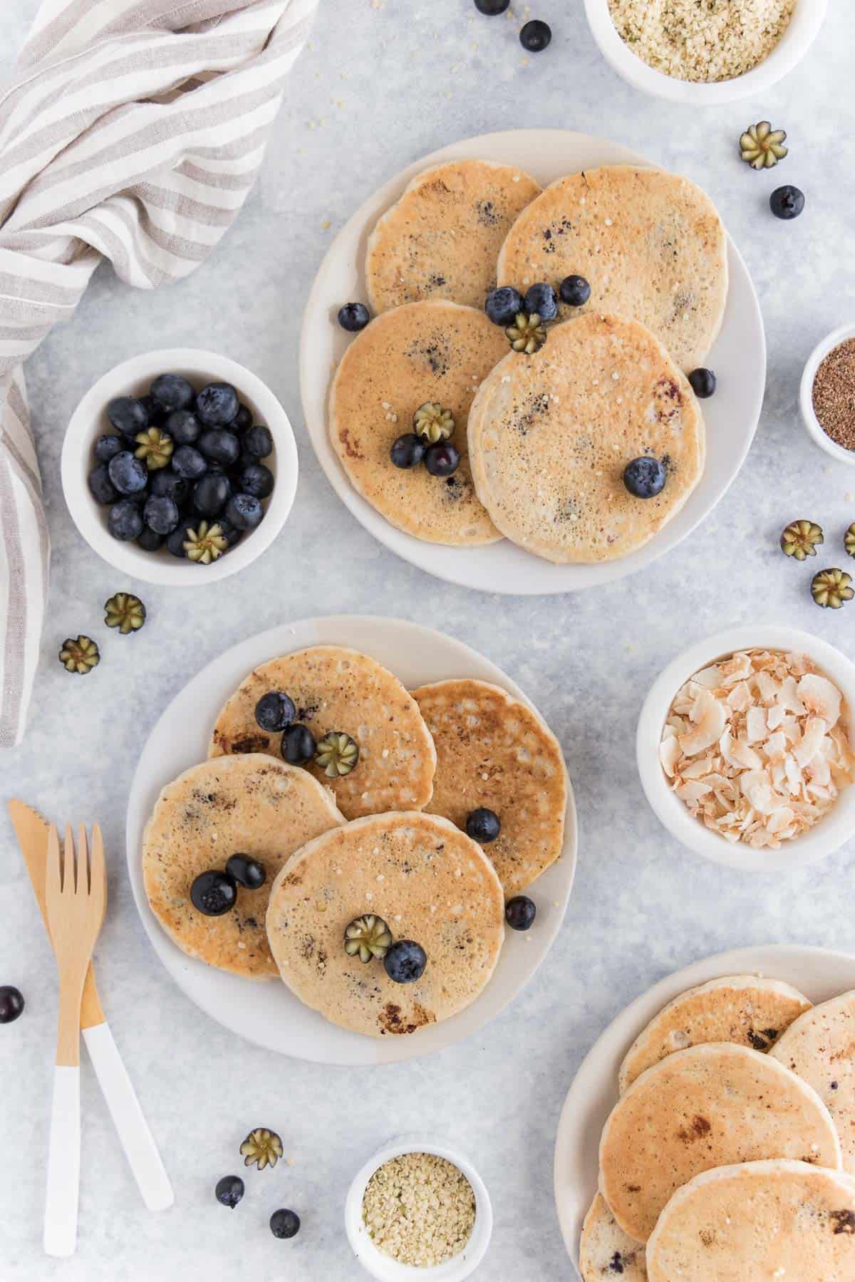pancakes laying on two plates surrounded by blueberries, hemp seeds, and coconut flakes