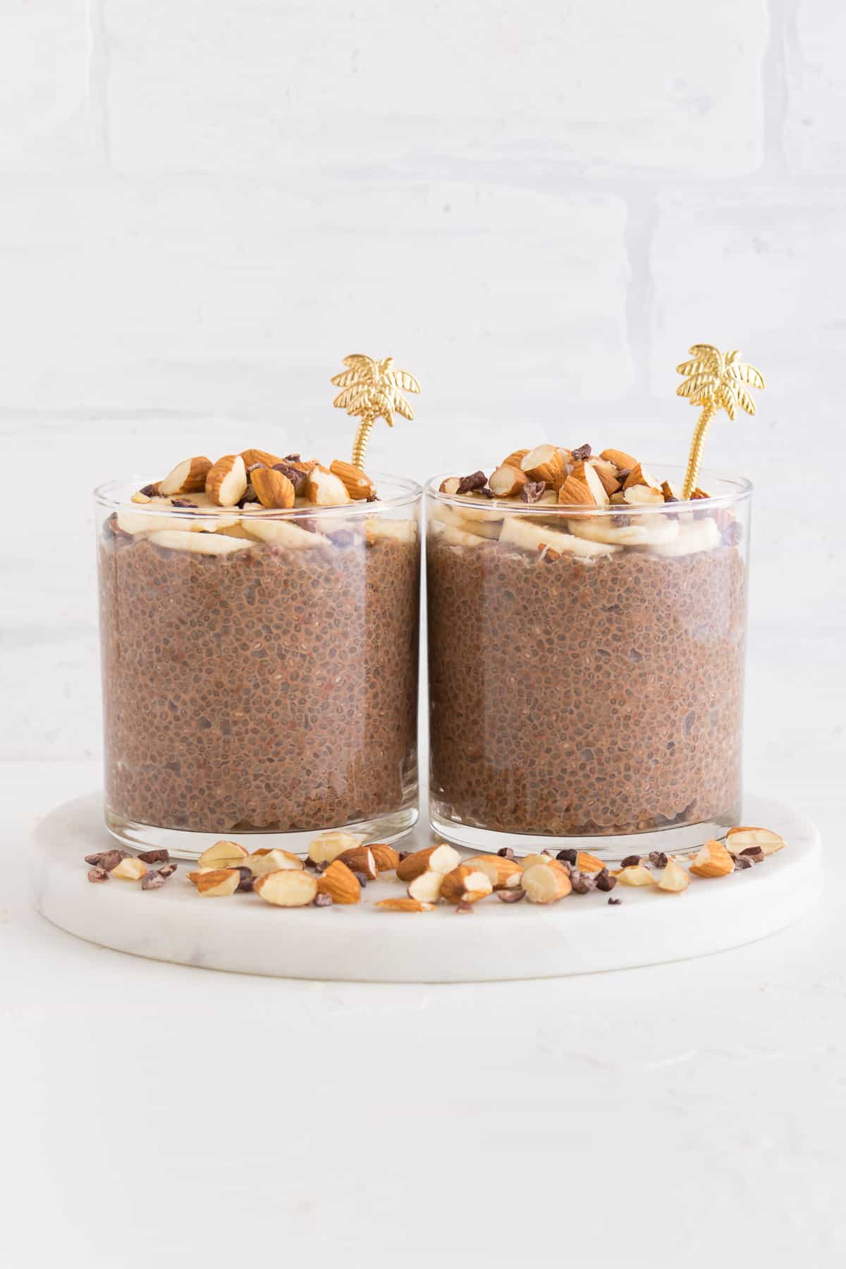 two glasses filled with chocolate chia seed pudding beside chopped almonds and cacao nibs
