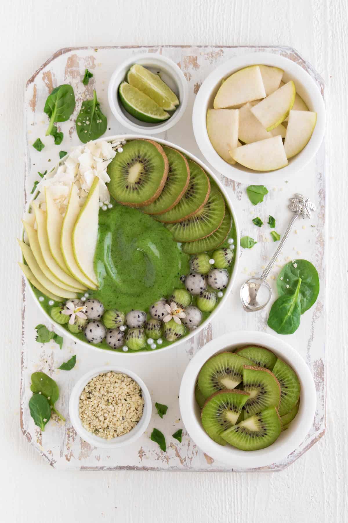 a white distressed cutting board with small bowls of hemp seeds, lime, kiwi, and pear