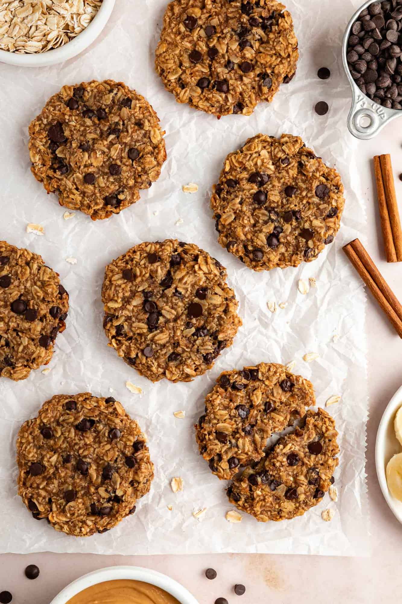 peanut butter chocolate chip banana oatmeal cookies laying on white parchment paper surrounded by ingredients in white bowls