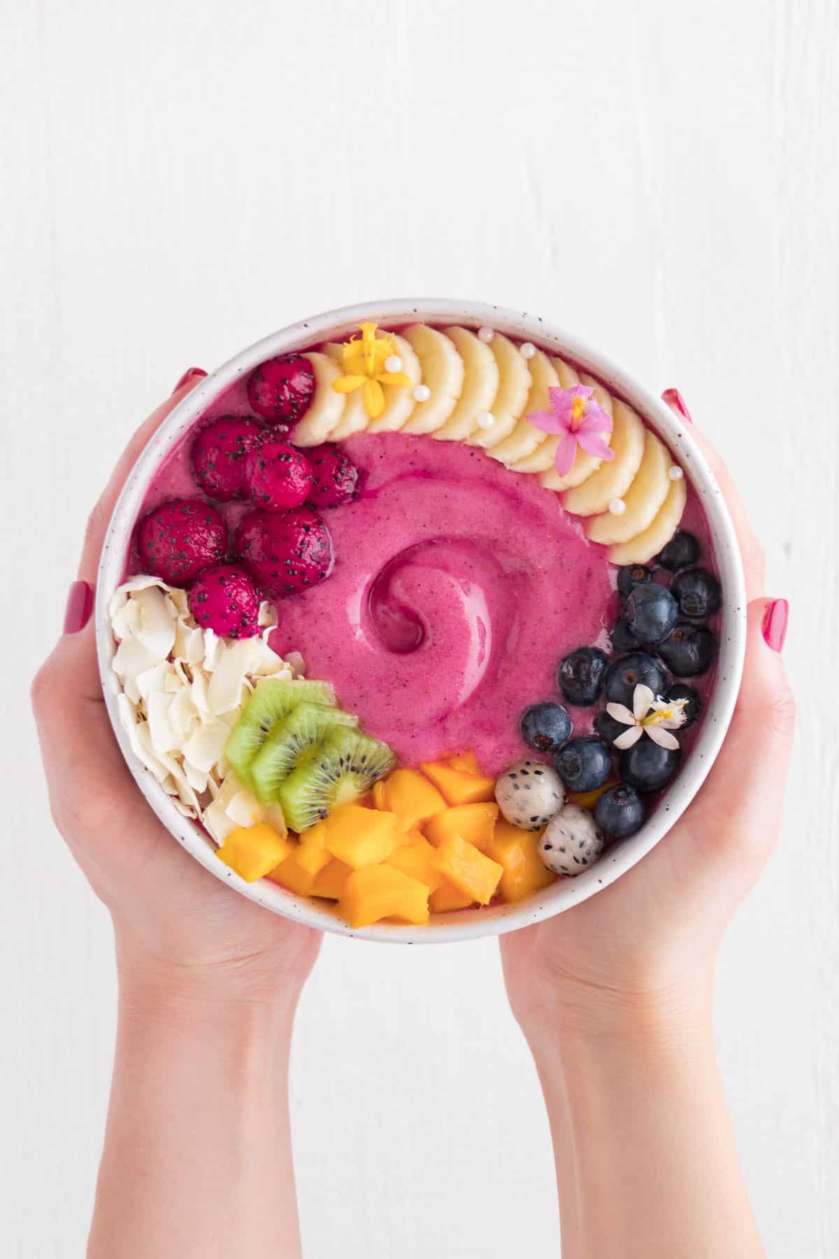 two hands holding a pink smoothie topped with with mango, kiwi, berries, banana, and coconut flakes inside a ceramic dish