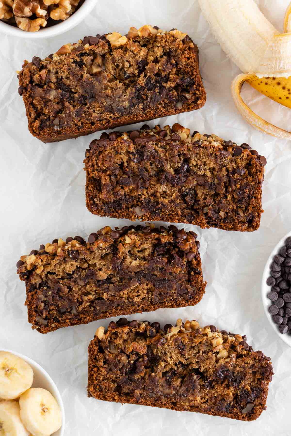 a hand grabbing a slice of vegan gluten-free banana bread on a white plate surrounded by almond milk, rolled oats, and walnuts