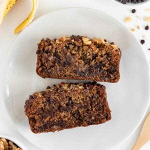 two slices of vegan gluten-free banana bread on a plate surrounded by a bowl of walnuts, a bowl of chocolate chips, and a peeled banana