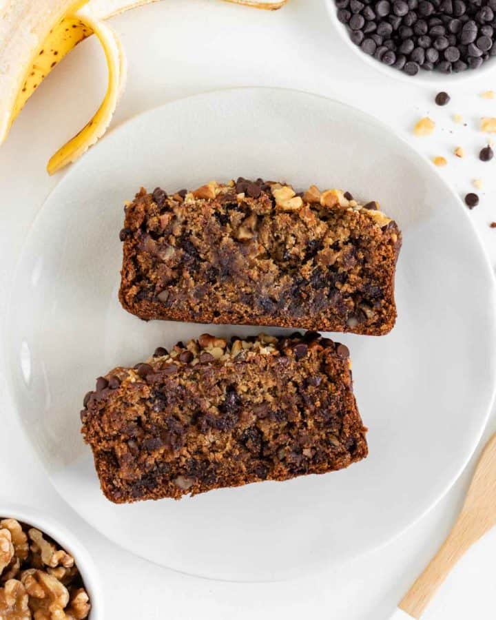 two slices of vegan gluten-free banana bread on a plate surrounded by a bowl of walnuts, a bowl of chocolate chips, and a peeled banana