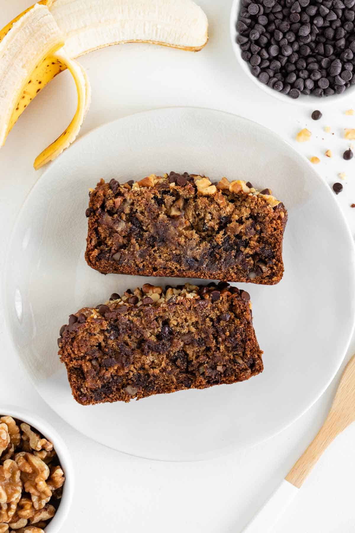 two slices of vegan gluten-free banana bread on a white plate surrounded by a bowl filled with walnuts, a bowl filled with oats, a glass of almond milk and a banana bbread loaf