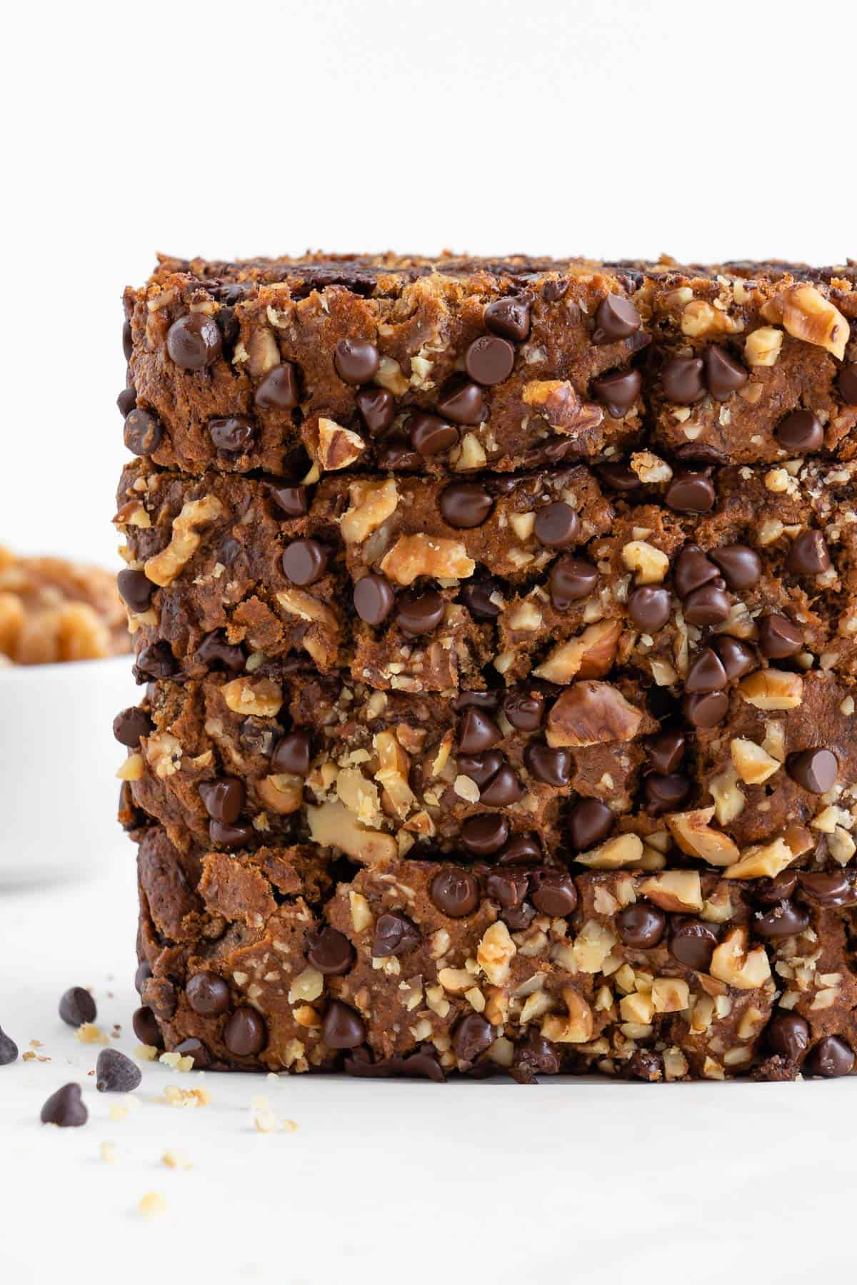 two slices of baked bread with chocolate chips beside a bowl of rolled oats, a bowl of walnuts, and a glass of almond milk