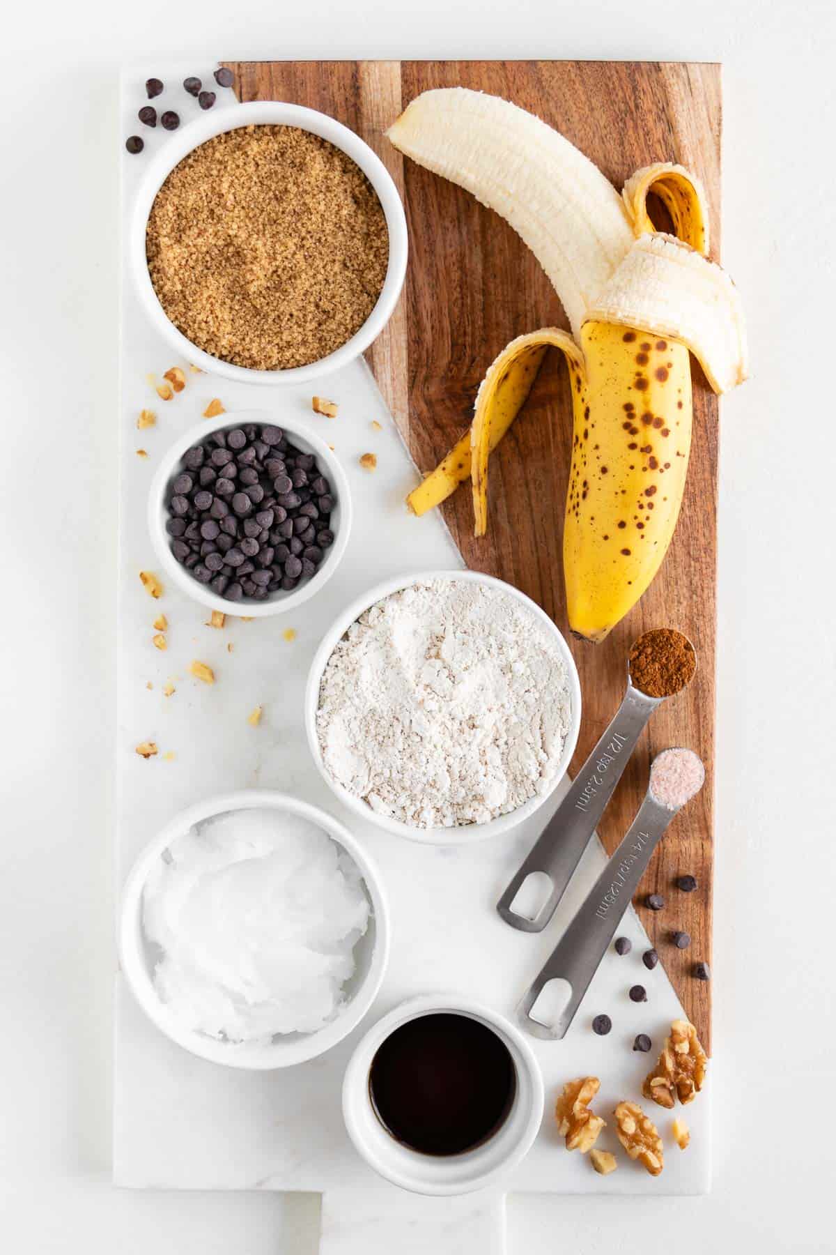 a marble and wooden board topped with bowls of brown sugar, chocolate chips, oat flour, coconut oil, vanilla extract, and a peeled banana