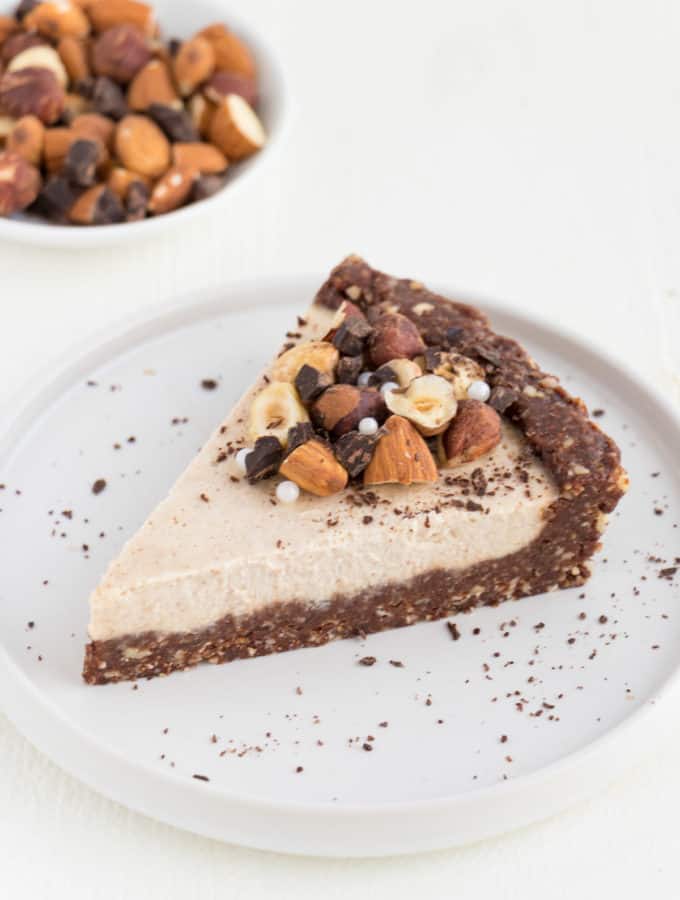 a slice of raw vegan cheesecake topped with almonds, hazelnuts, and chocolate