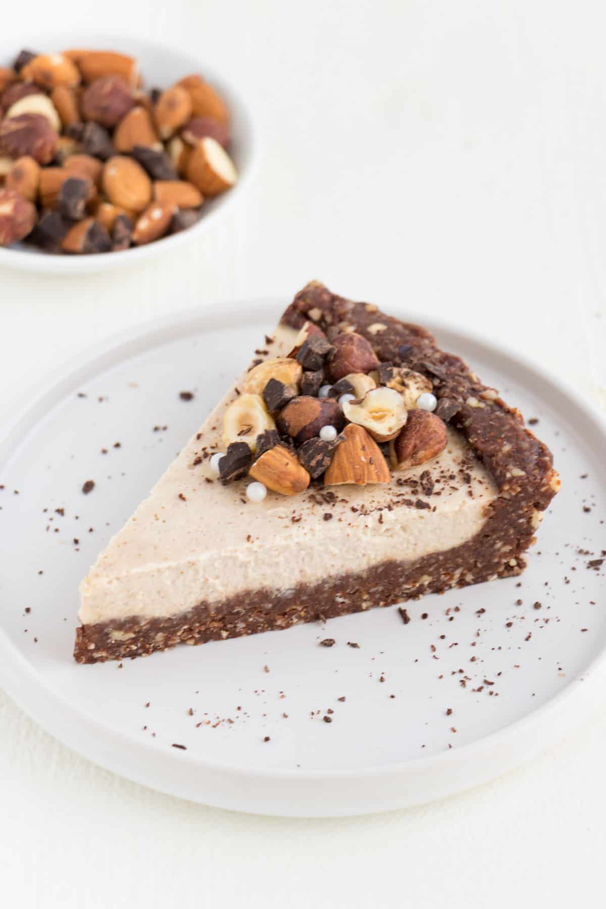 a slice of raw vegan cheesecake topped with almonds, hazelnuts, and chocolate