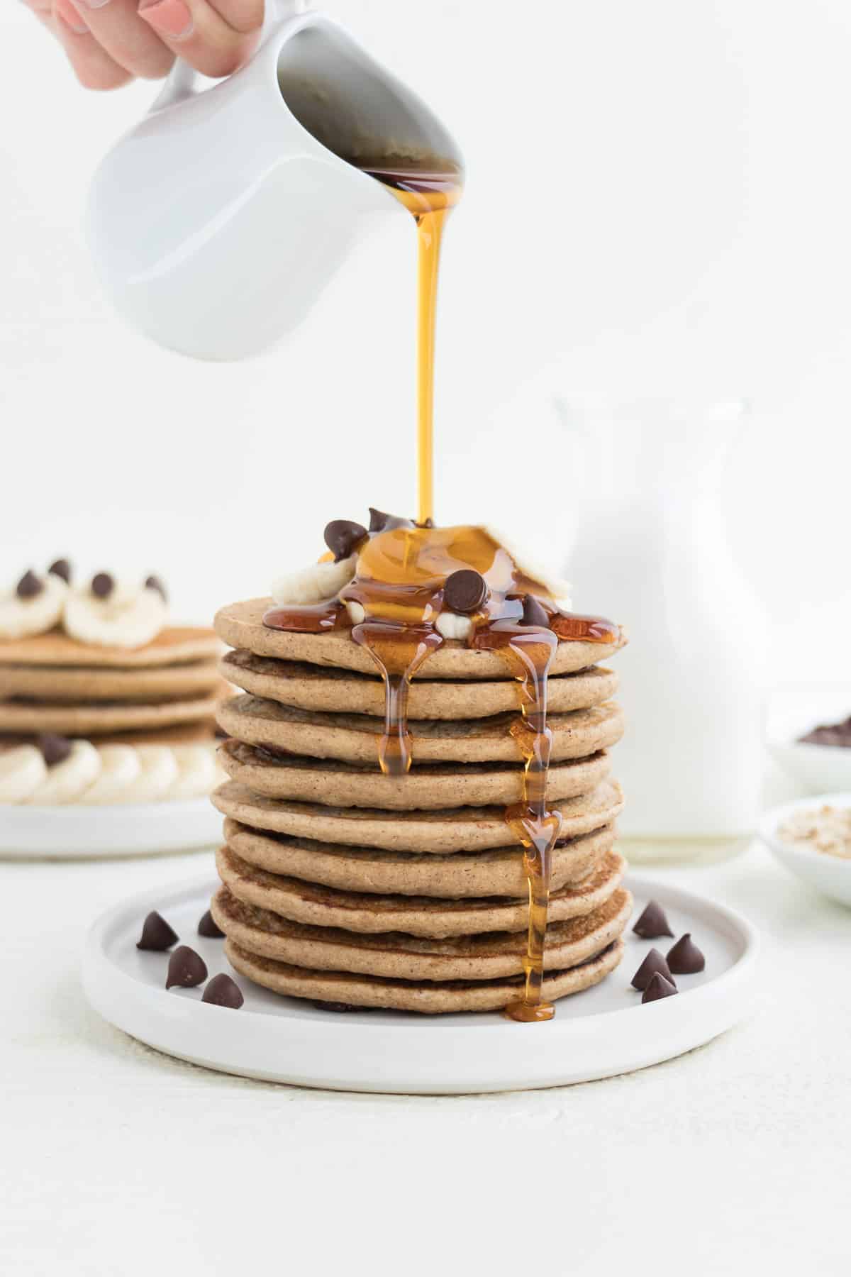 pouring maple syrup over a stack of vegan chocolate chip banana pancakes