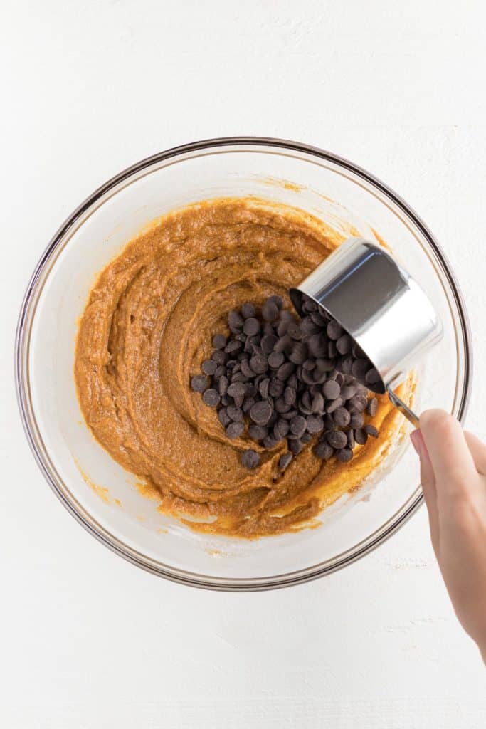 a hand pouring a cup of chocolate chips into pumpkin bar batter