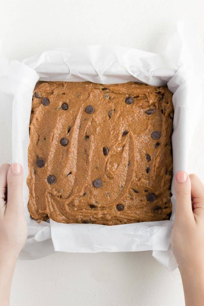 two hands holding a square pan filled with chocolate chip pumpkin bar batter