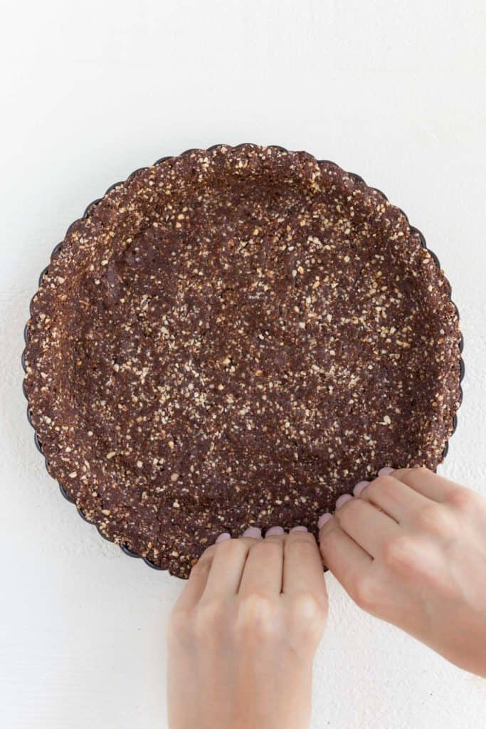 two hands pressing a no bake chocolate almond crust into a springform tart pan
