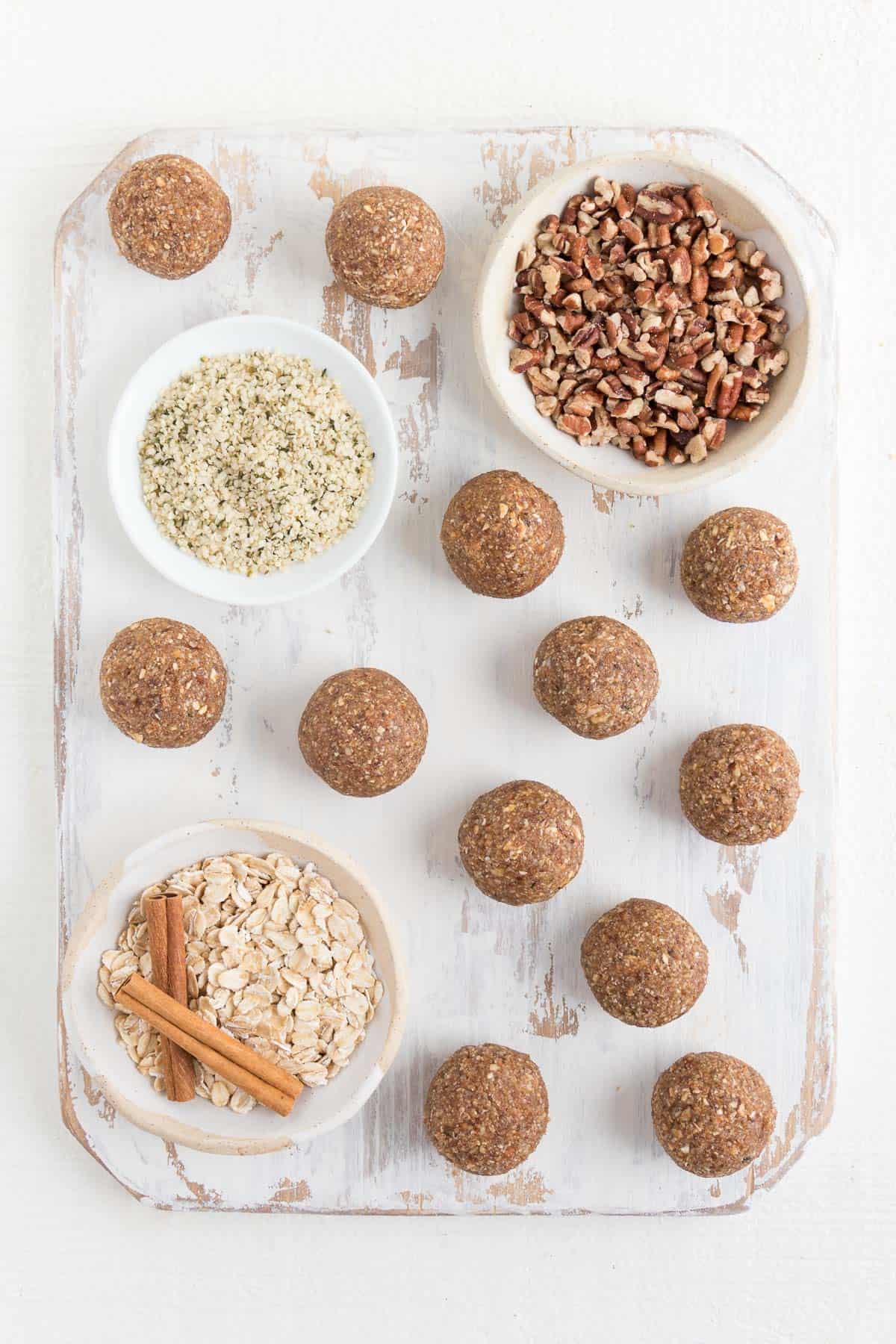 no-bake pecan balls on a white distressed cutting board with small bowls of cinnamon, oats, pecans, and hemp seeds