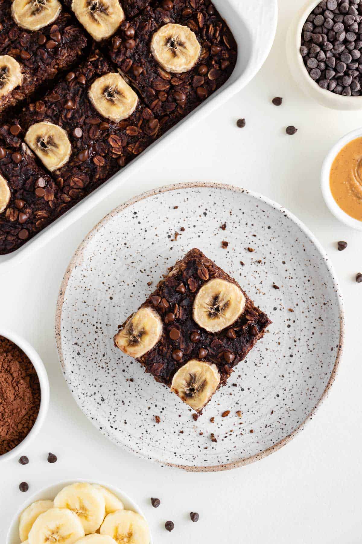 a slice of chocolate baked oatmeal on a speckled ceramic plate surrounded by peanut butter, cacao, and a tray of oatmeal bake