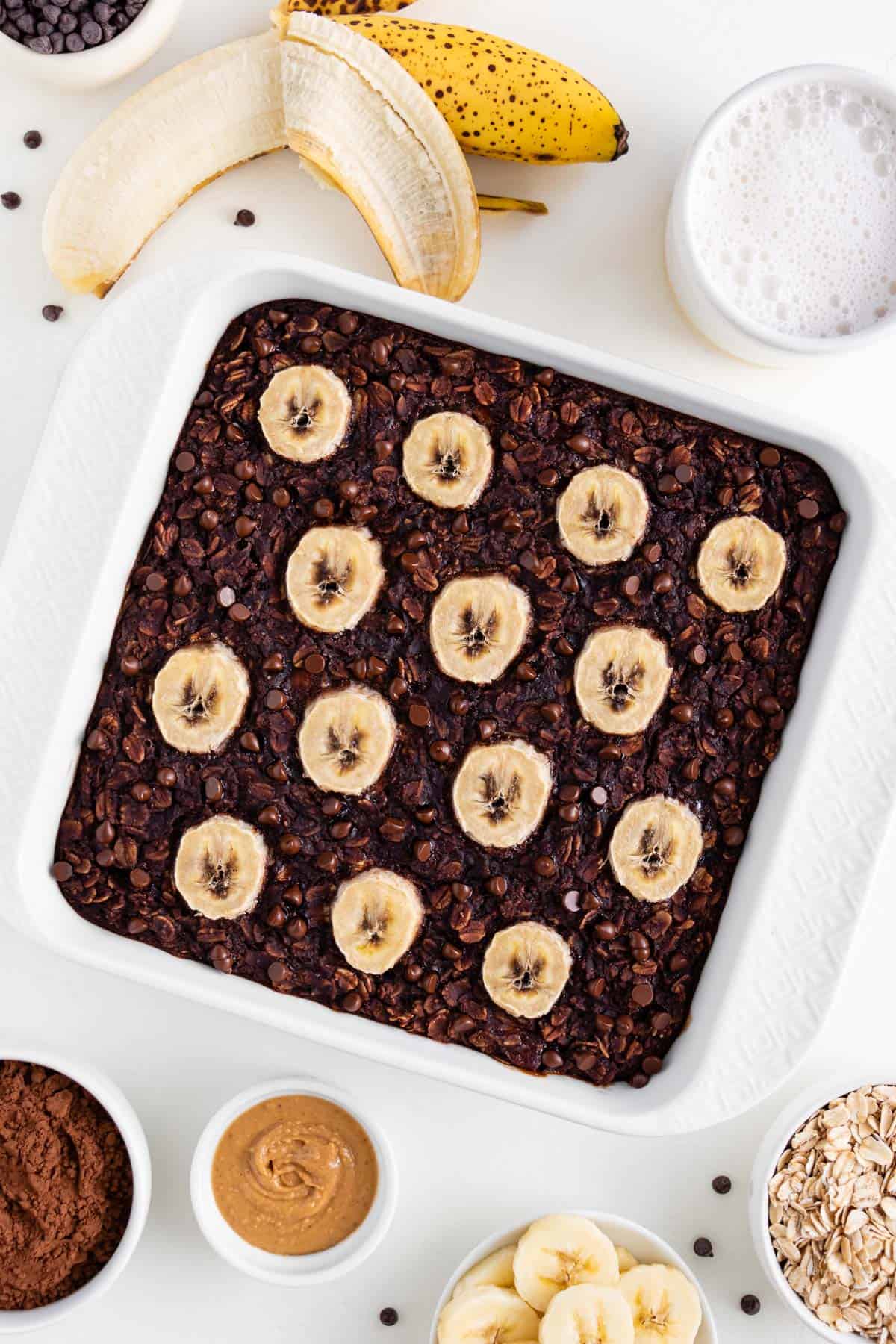 overhead image of chocolate banana baked oatmeal inside a white square baking dish, surrounded by a peeled banana, glass of almond milk, and bowls of ingredients