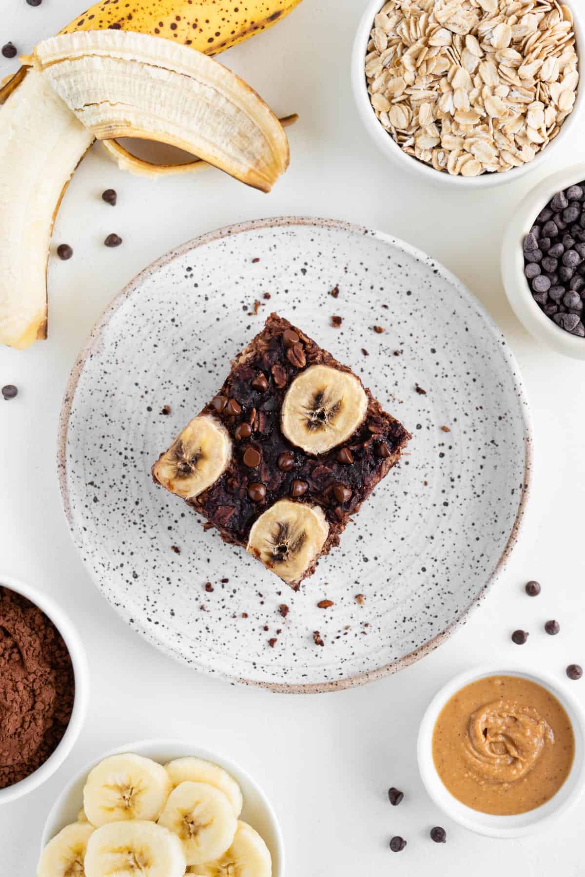 a square of chocolate baked oatmeal on a speckled ceramic plate surrounded by a peeled banana, and bowls containing rolled oats, peanut butter, cacao, and chocolate chips