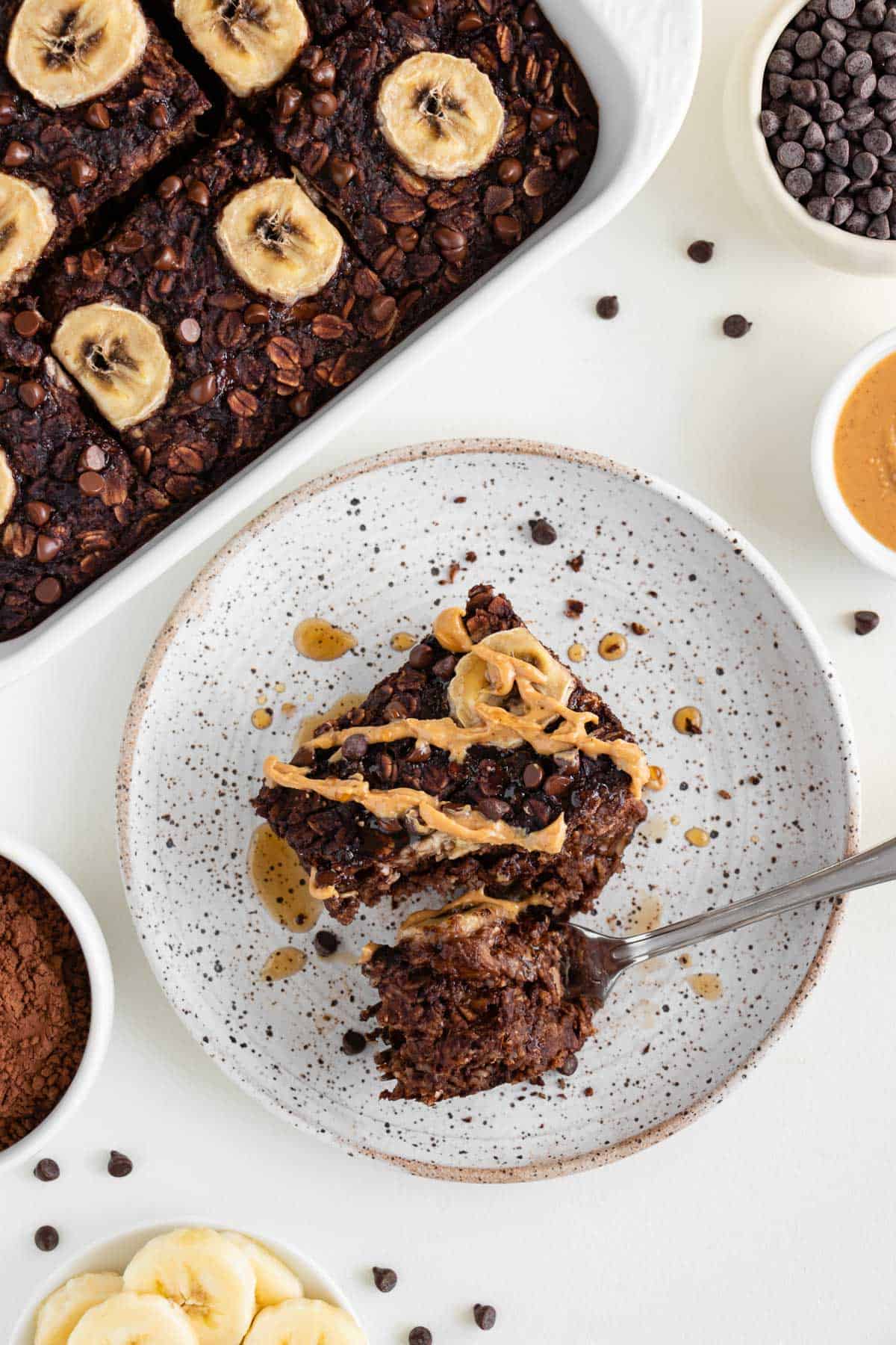 a tray of chocolate banana baked oatmeal placed beside a partially eaten slice that's topped with peanut butter, maple syrup, and sliced bananas