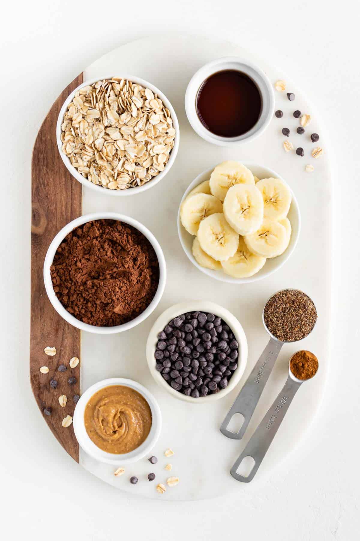 a marble board topped with small bowls containing rolled oats, cacao, bananas, peanut butter, chocolate chips, vanilla, cinnamon, and ground flaxseed