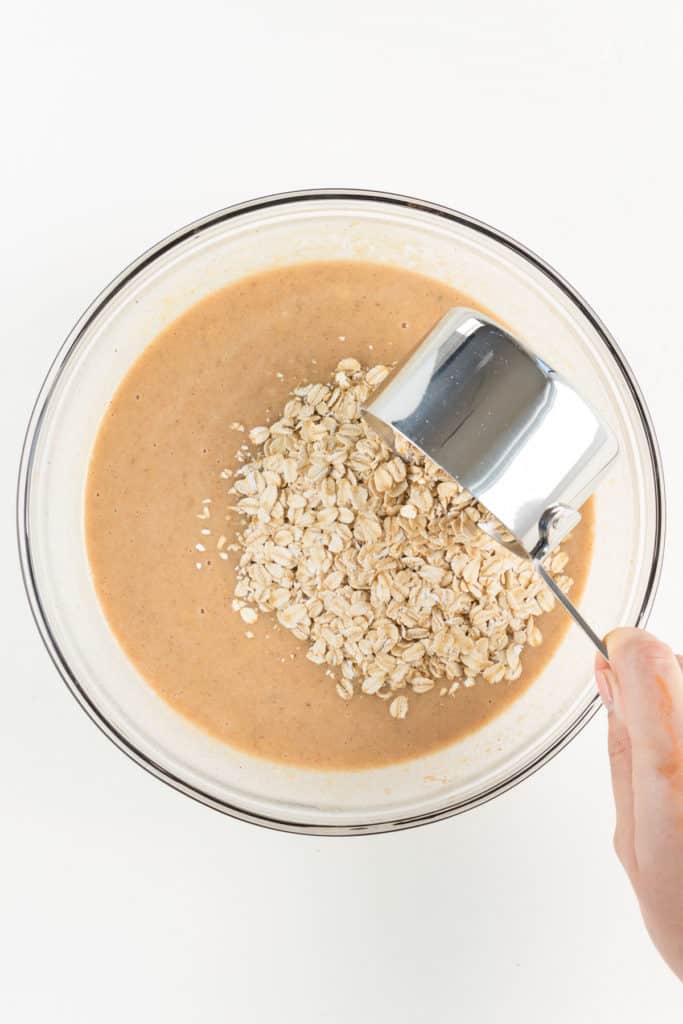 hand holding a metal measuring cup pouring rolled oats into a brown batter