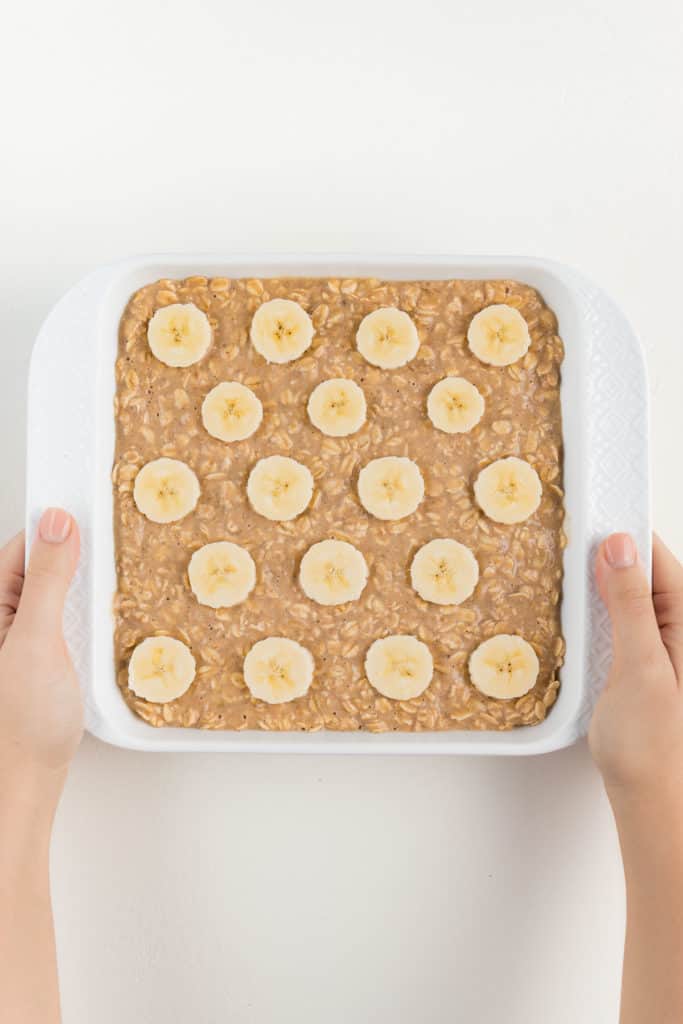 two hands holding a white square baking dish that contains oatmeal batter with bananas