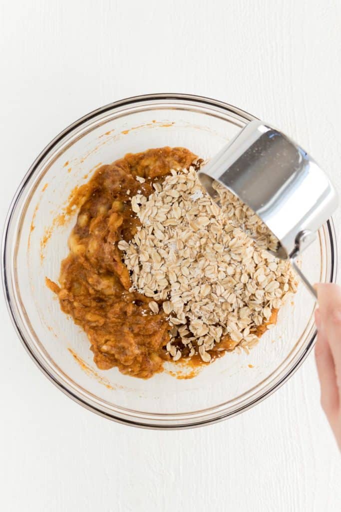 hand pouring a metal measuring cup of rolled oats into pumpkin batter