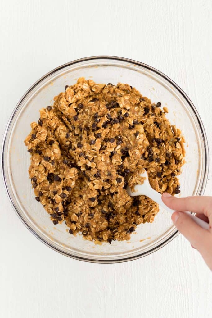 hand holding a white spatula mixing together pumpkin chocolate chip oatmeal cookie batter inside a glass bowl