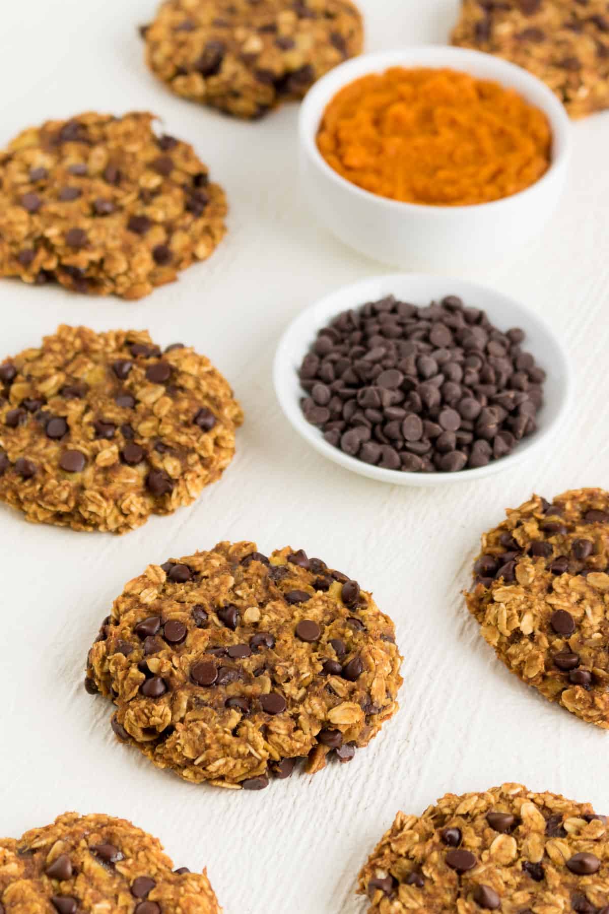 two small white bowls containing pumpkin puree and mini chocolate chips surrounded by chocolate chip oatmeal cookies