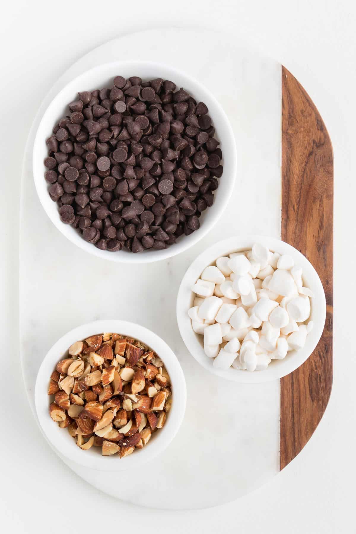 three white bowls containing vegan chocolate chips, roasted almonds, and mini marshmallows on top of a white marble board