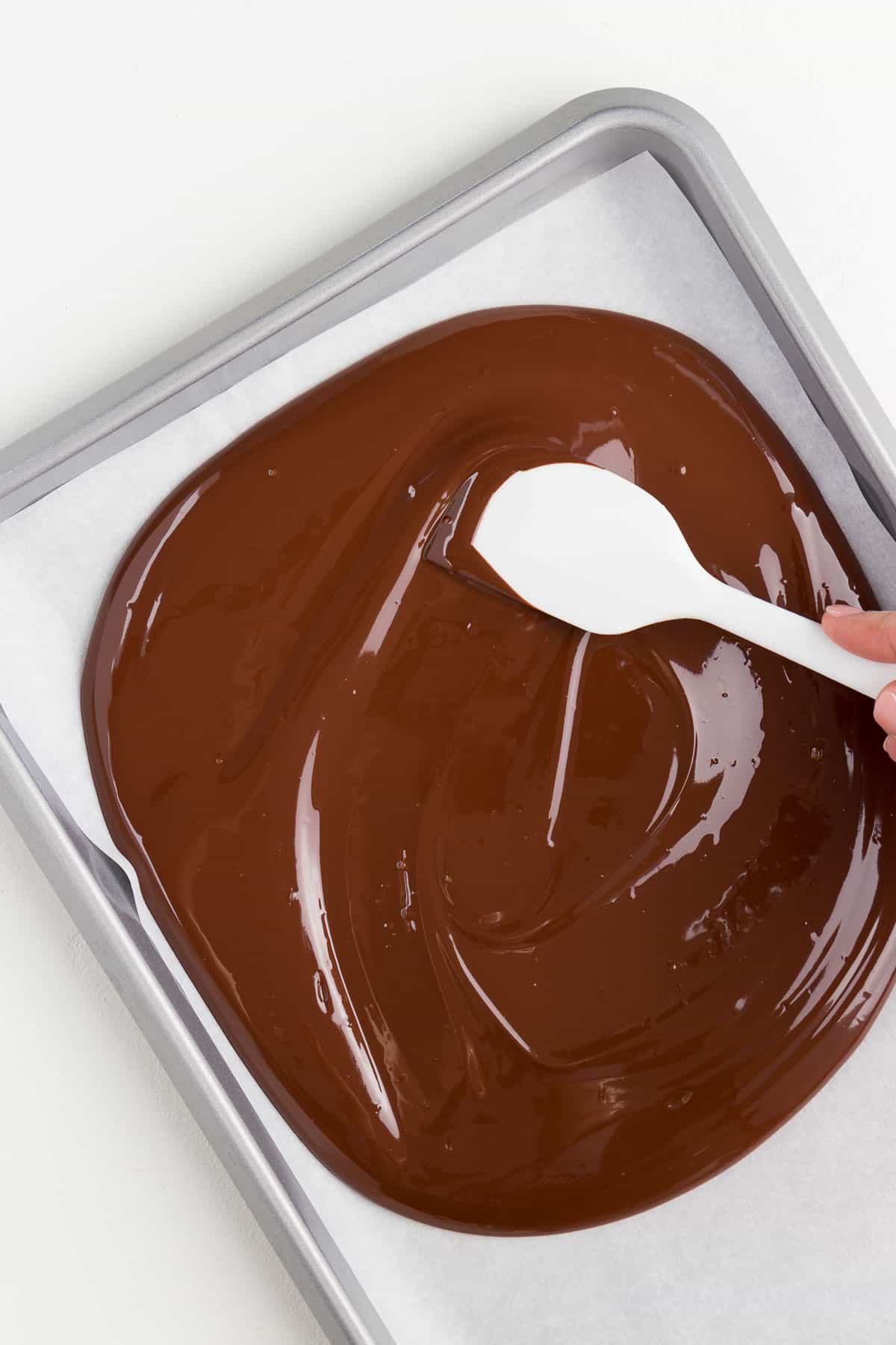 hand holding a white spatula spreading melted chocolate across a silver baking sheet