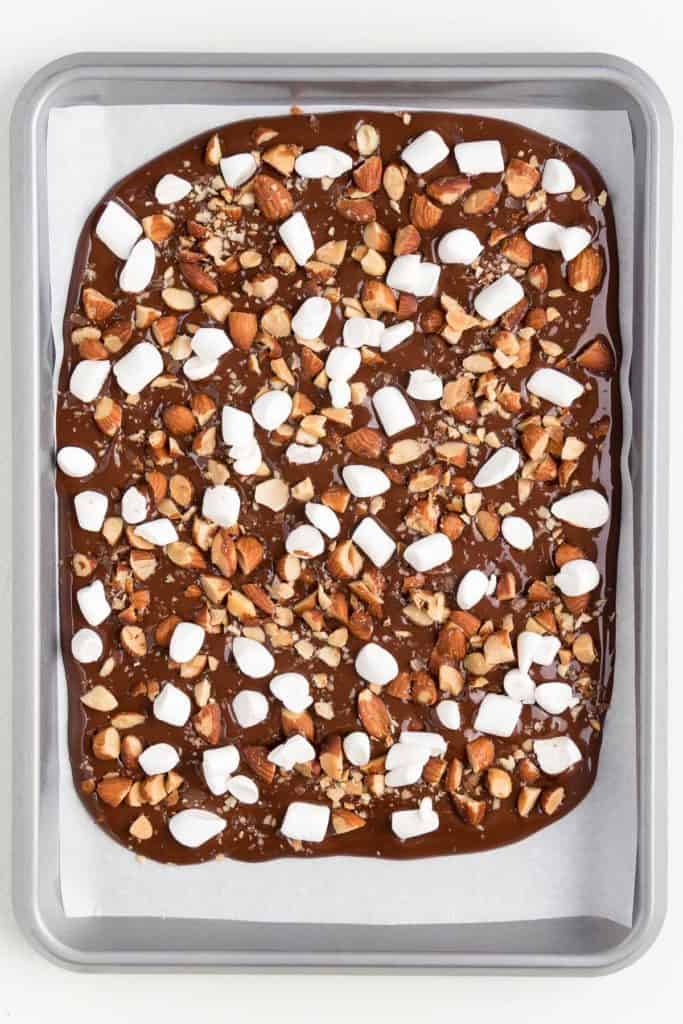 melted chocolate spread across a silver baking sheet topped with chopped almonds and mini marshmallows