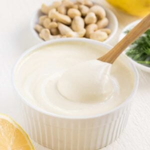 a wooden spoon scooping vegan cashew sour cream out of a white ramekin surrounded by lemons, dill, and cashews