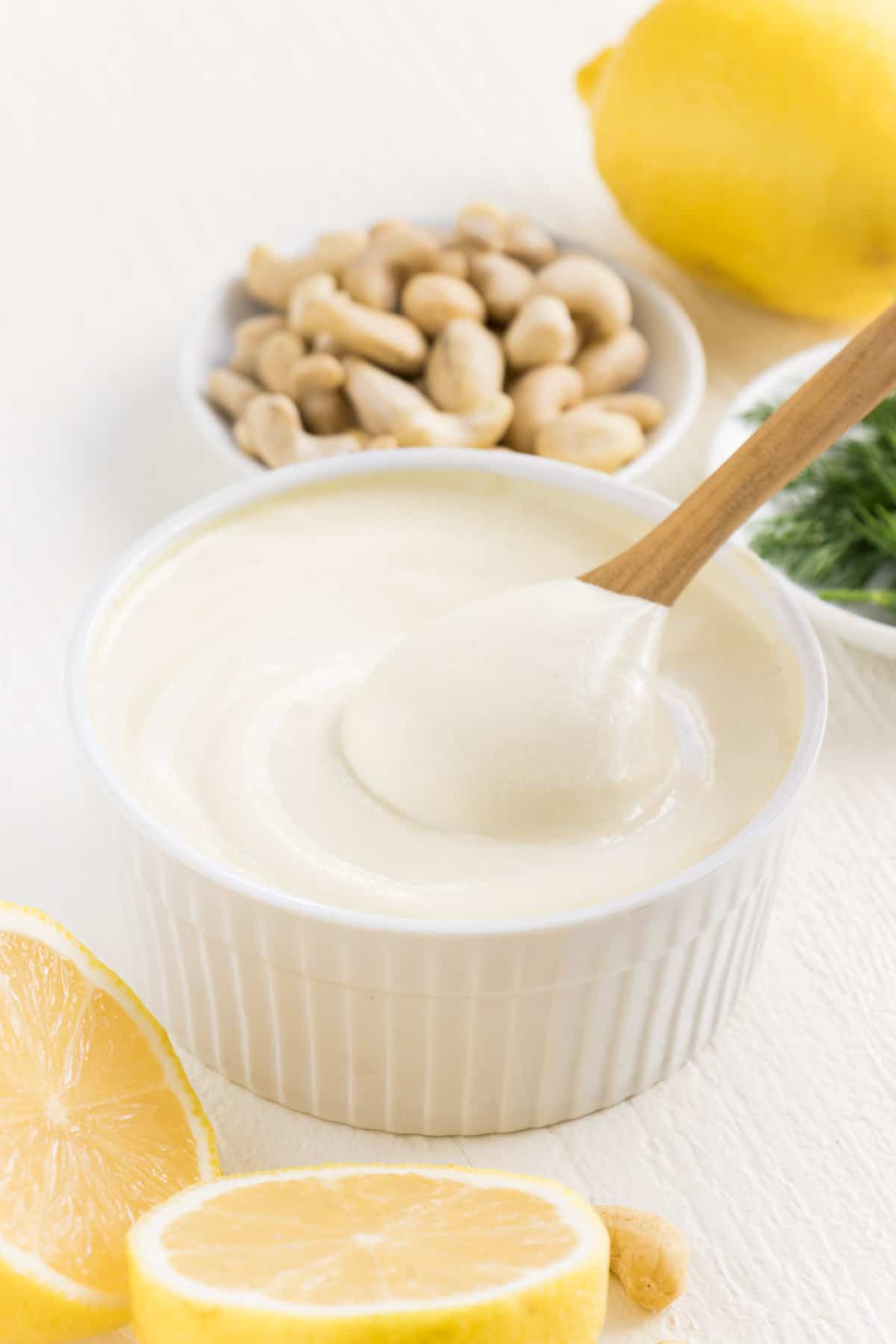 a wooden spoon scooping vegan cashew sour cream out of a white ramekin surrounded by lemons, dill, and cashews