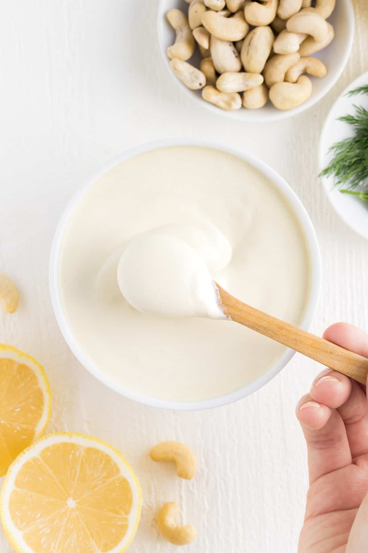 a hand holding a wooden spoon scooping vegan cashew sour cream out of a white bowl