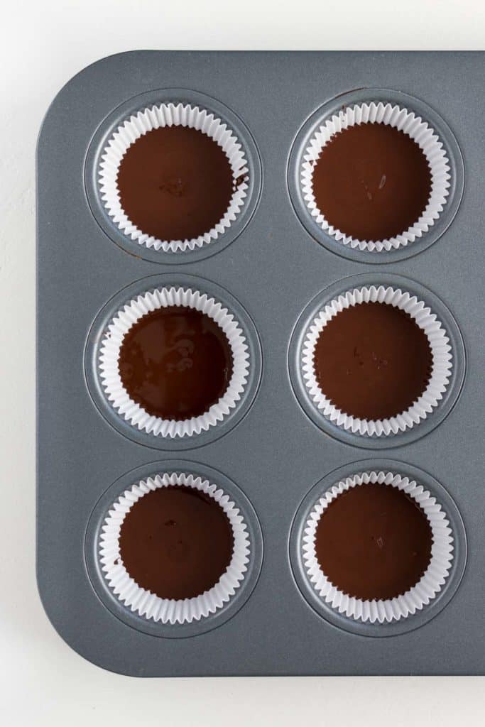 a metal muffin tin filled with white muffin liners each containing melted vegan chocolate