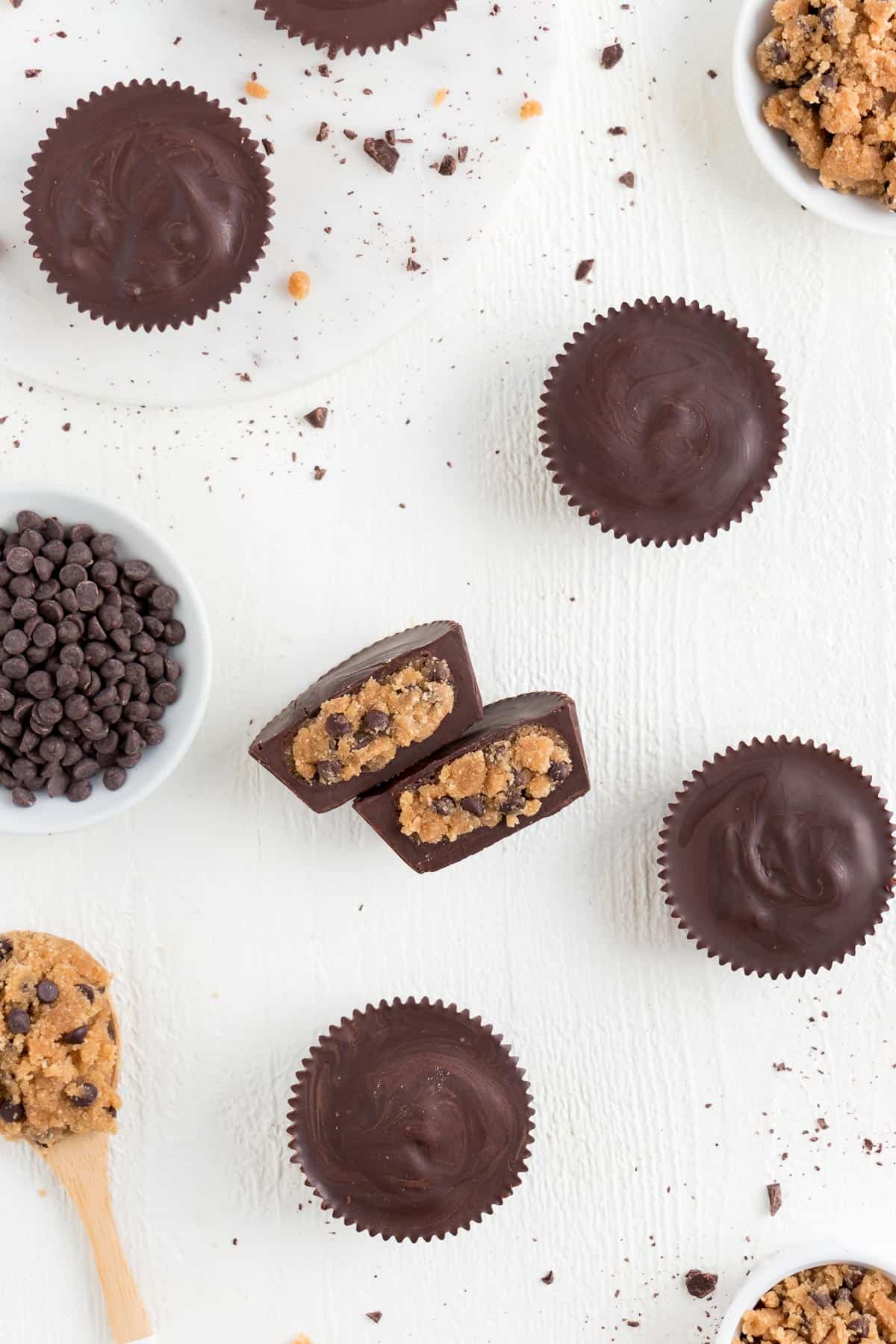 vegan chocolate chip cookie dough cups surrounded by a wooden spoon with cookie dough, chocolate chips, and chocolate shavings