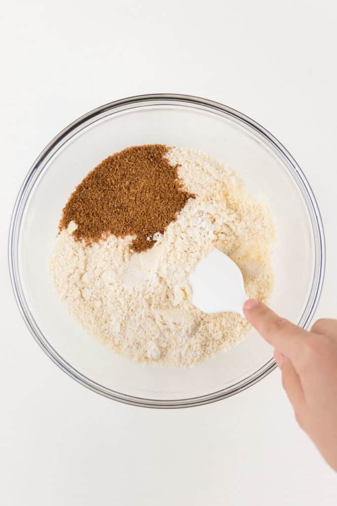 hand holding a white spatula mixing almond flour, coconut flour, and coconut sugar in a glass pyrex bowl