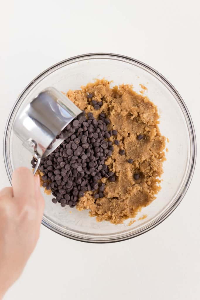 hand pouring a metal measuring cup containing chocolate into a glass bowl filled with cookie dough