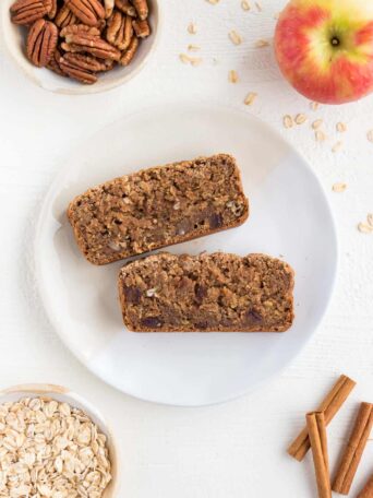 two slices of apple cinnamon bread on a white ceramic plate surrounded by a honeycrisp apple, cinnamon sticks, rolled oats, and pecans
