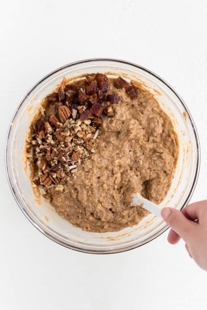a hand holding a white spatula mixing batter with crushed pecans and chopped medjool dates