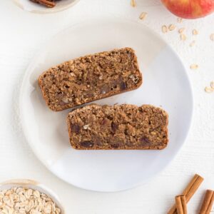 two slices of apple cinnamon bread on a white plate surrounded by cinnamon sticks, pecans, oats, and honeycrisp apple