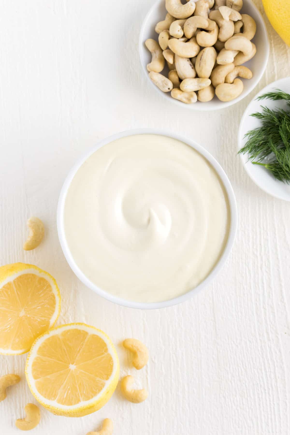 sour cream inside a white bowl surrounded by lemons, cashews, and dill