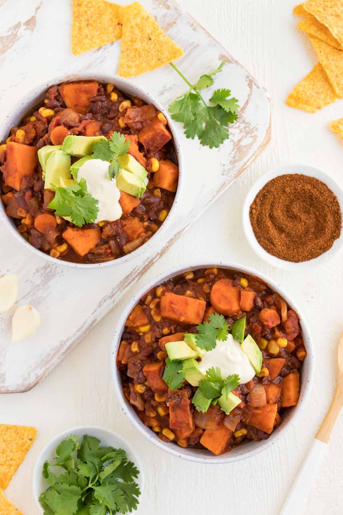 two bowls of chili on a distressed white cutting board with a small bowl of chili powder, a small bowl of cilantro, and tortilla chips