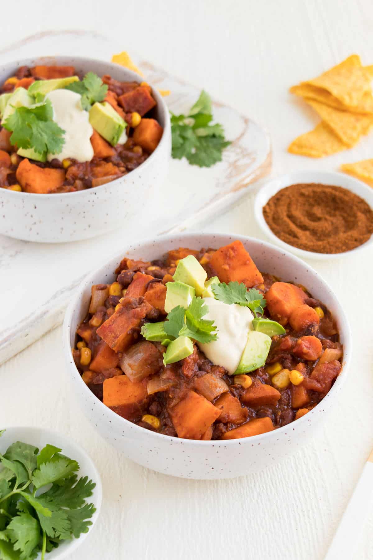 a white ceramic bowl filled with vegan sweet potato black bean chili surrounded by cilantro, tortilla chips, and chili powder