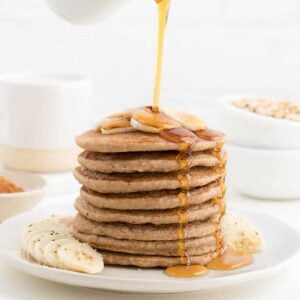 hand pouring maple syrup over a stack of banana pancakes