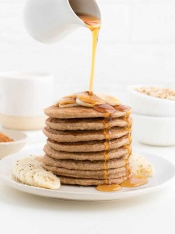 hand pouring maple syrup over a stack of banana pancakes