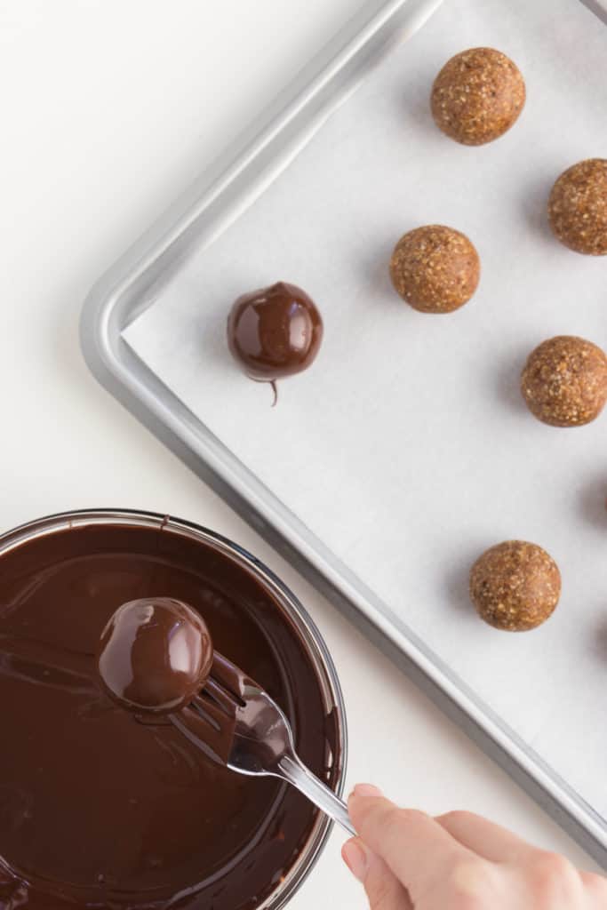 metal fork dipping a gingerbread truffle into a glass bowl of melted chocolate, next to a baking sheet with gingerbread truffles