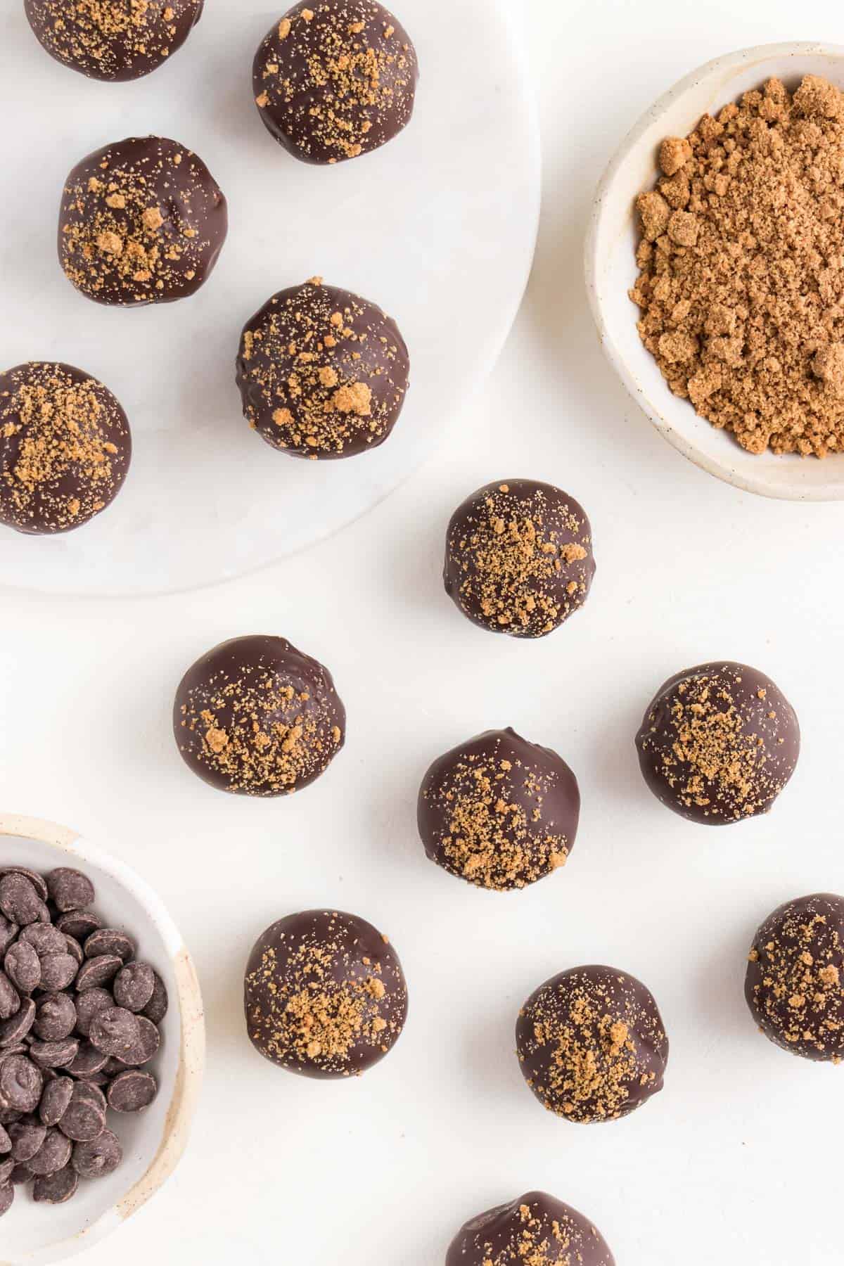 chocolate gingerbread truffles scattered across a white surface beside a small ceramic bowl filled with chocolate chips and crumbled gingersnap cookies