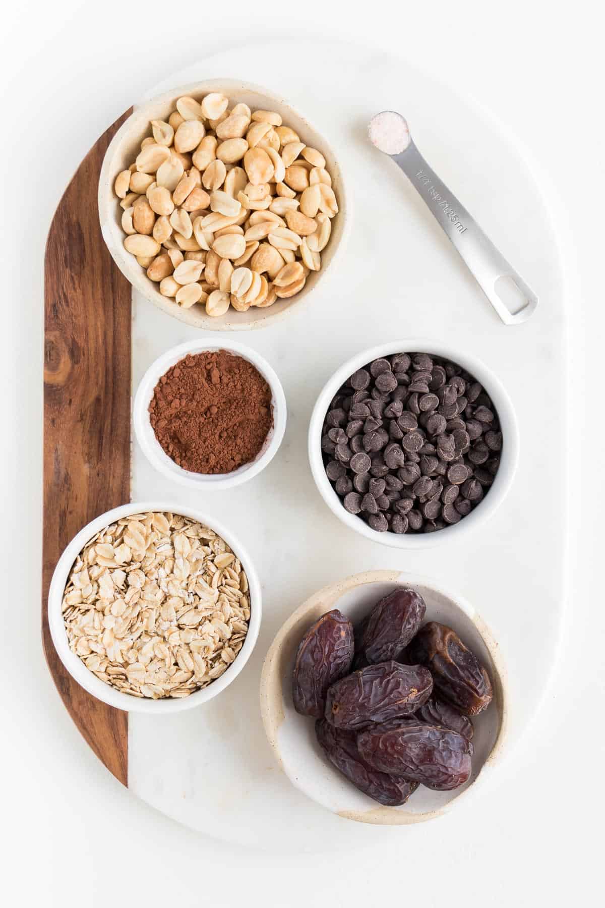 a marble and wood cutting board topped with small bowls containing chopped peanuts, cocoa powder, chocolate chips, oats, medjool dates, and a metal teaspoon
