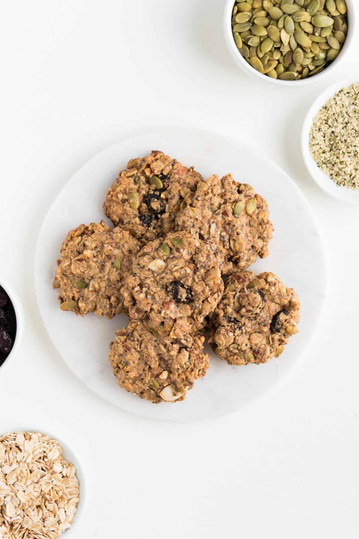 six superfood breakfast cookies surrounded by small white bowls filled with pumpkin seeds, cranberries, rolled oats, and hemp seeds